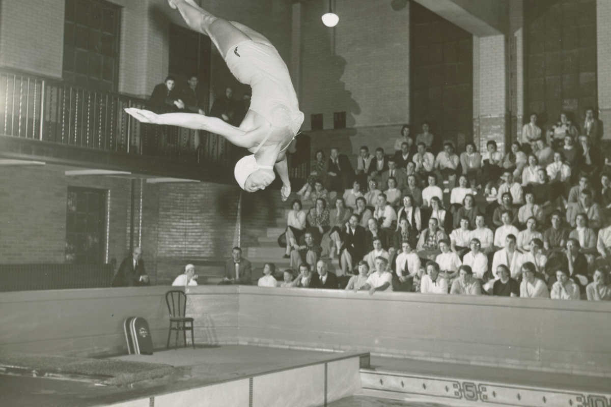A person diving into an indoor pool in front of a crowd on bleachers