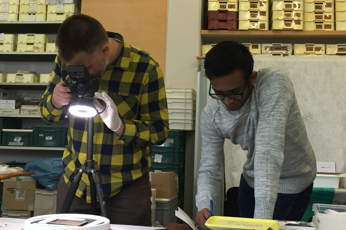 Professor wearing a yellow and black plaid shirt uses a ring light on a DSLR camera to image a wooden tablet sitting on the table. Undergraduate student in a grey sweatshirt and glasses is standing next to the professor taking notes next to him.