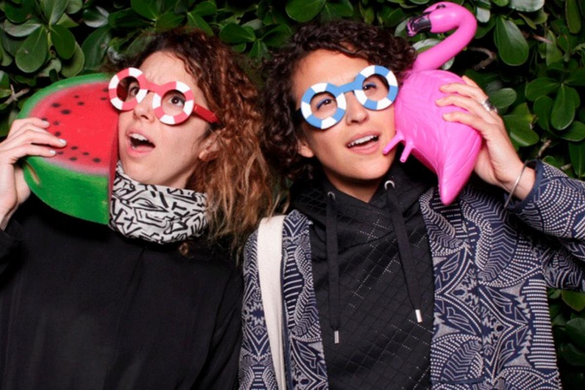 Ece Tankal and Carmen Aguilar y Wedge. They are wearing fun glasses and holding a plastic flamingo and watermelon.