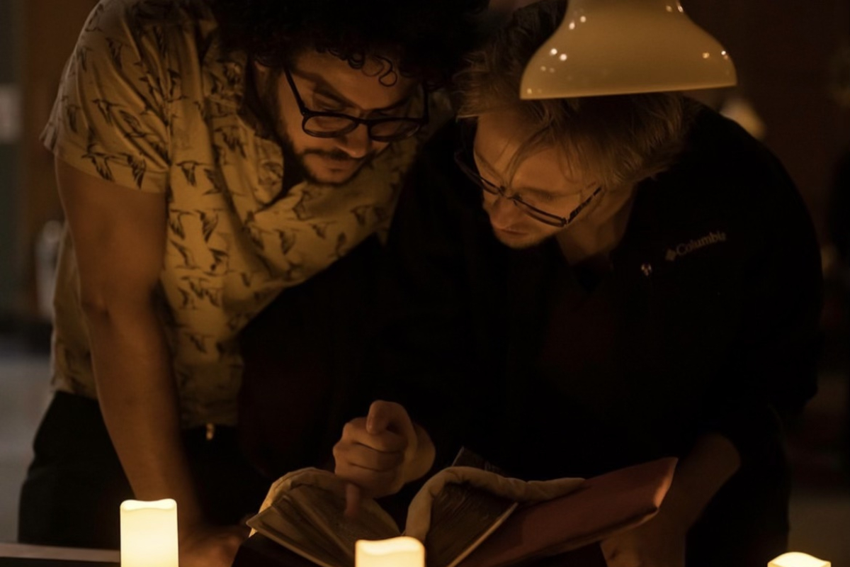 Two people bend over a medieval manuscript on a table, surrounded by electric candles. They gesture at the manuscript and look at it closely.