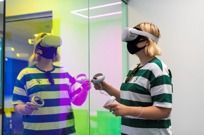 A woman using a VR headset; her reflection is visible in the glass wall she's standing next to