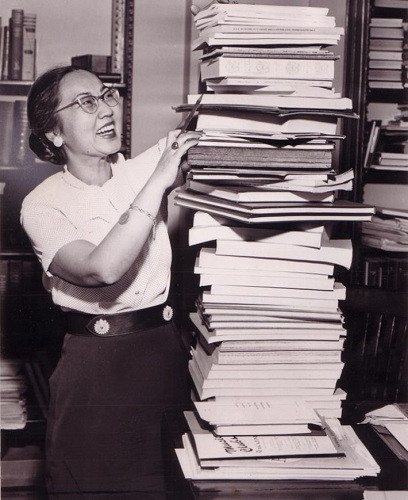 Ruth Watanabe standing near a tall stack of publications