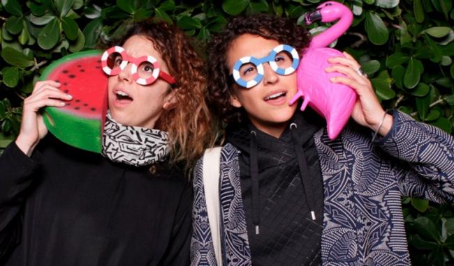 Ece Tankal and Carmen Aguilar y Wedge. They are wearing fun glasses and holding a plastic flamingo and watermelon.