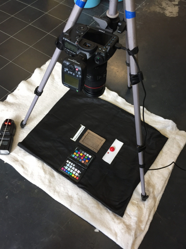 Reflectance Transformation Imagining (RTI) set-up shows a DSLR camera on a tripod with the camera lens pointing down toward the ground. There is a cloth on the ground with a black board on it. On the board is the RTI set-up which includes the wooden tablet being imaged, color bars, a ruler and small red targets.