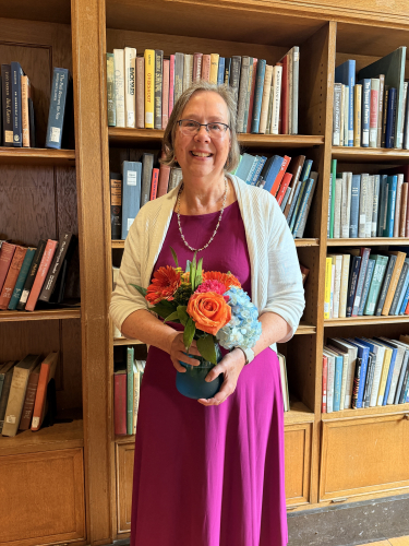 Photo of Sue Cardinal standing in front of a bookshelf holding flowers