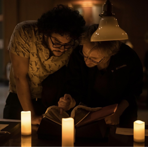 Two people bend over a medieval manuscript on a table, surrounded by electric candles. They gesture at the manuscript and look at it closely.