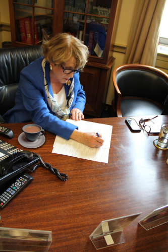 Congresswoman Louise Slaughter at a desk writing on a document with a cup of tea next to her