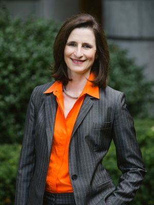Photo of Mary Ann Mavrinac standing in a grey blazer and orange shirt